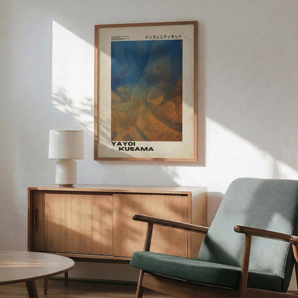 May include: Framed poster with abstract dotted pattern in blue, orange and brown tones displayed above wooden sideboard in modern living room. Green upholstered mid-century chair with wooden arms sits nearby, white ceramic table lamp on credenza, sunlight streaming across white walls creating geometric shadows.