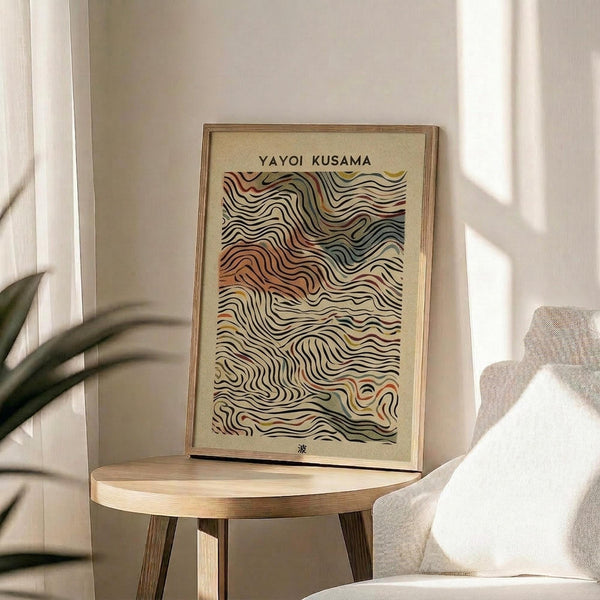 May include: Framed art print with flowing wave patterns in earth tones of orange, brown, yellow and cream displayed on a round wooden side table next to a beige upholstered chair. Natural sunlight streams across the neutral-toned room setting with a plant visible in the corner.