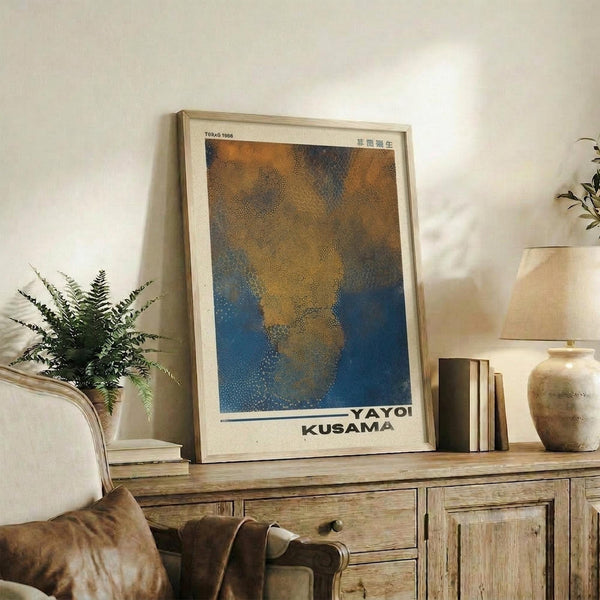 May include: Framed art poster with abstract dotted pattern in orange, brown, and blue tones displayed on rustic wooden sideboard. Natural wood frame surrounds the artwork which features organic flowing shapes created with small dots. Living room setting includes cream upholstered chair, fern plant, ceramic vase, and stacked books on weathered wood furniture against white wall.