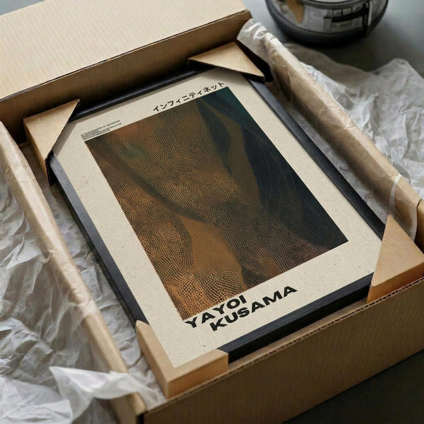 May include: Framed art print featuring abstract dotted pattern in earth tones of brown, green and orange, displayed in black frame on cream mat. Print sits in opened cardboard shipping box with protective wrapping materials and wooden frame corners visible. Minimalist unboxing setup on white surface.
