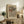May include: Framed poster with flowing wave-like patterns in earth tones of rust, sage green, cream, and brown against a beige background, displayed on a rustic wooden sideboard. Scene shows the artwork leaning against a white wall beside a fern plant, stacked books, and ceramic vases in a warm, natural living room setting.