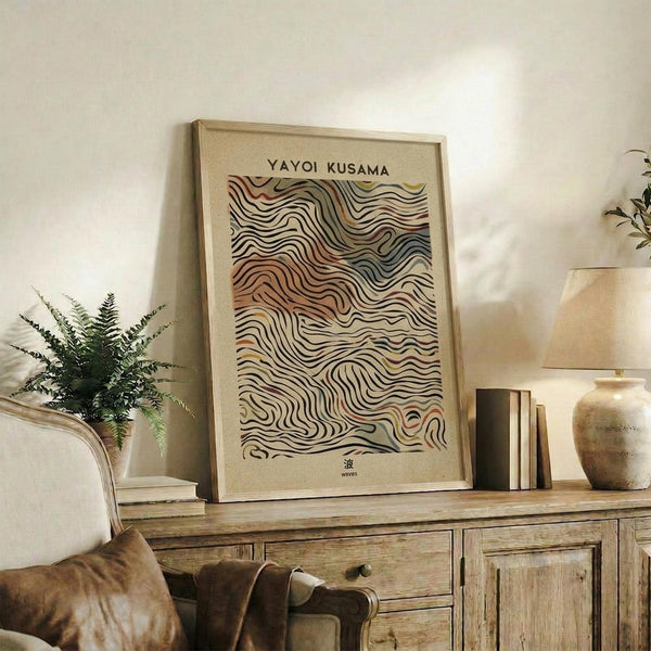 May include: Framed poster with flowing wave-like patterns in earth tones of rust, sage green, cream, and brown against a beige background, displayed on a rustic wooden sideboard. Scene shows the artwork leaning against a white wall beside a fern plant, stacked books, and ceramic vases in a warm, natural living room setting.