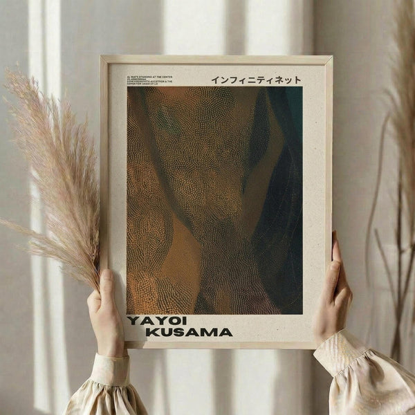 May include: Person holding framed poster print featuring abstract dotted pattern in earthy brown, orange and dark green tones, styled after Kusama's infinity net artwork. Light wood frame against neutral beige background with dried pampas grass stems. Japanese text visible at top of print.