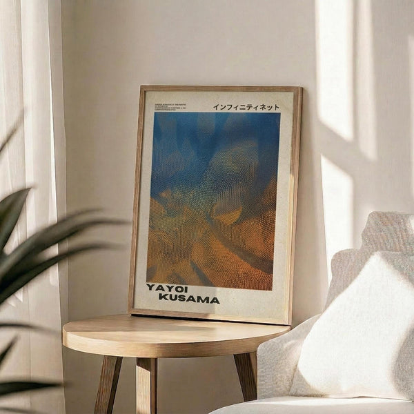 May include: Framed poster with dotted blue and orange abstract landscape artwork displayed on round wooden side table next to beige upholstered chair and plant. Natural lighting creates soft shadows on white wall. Light wood frame complements minimalist Scandinavian-style interior design with neutral tones.