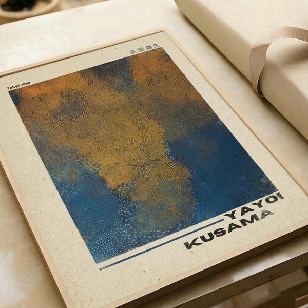 A vintage-style poster print featuring an abstract dotted pattern in golden yellow and deep blue tones, displayed on cream-colored paper with "YAYOI KUSAMA" text at the bottom and "Tokyo 1998" in the corner. The artwork shows Kusama's signature infinity net style with organic, flowing forms created through small dots. The print lies flat on a neutral surface next to rolled paper tubes, showing the poster's actual size and cream paper texture.