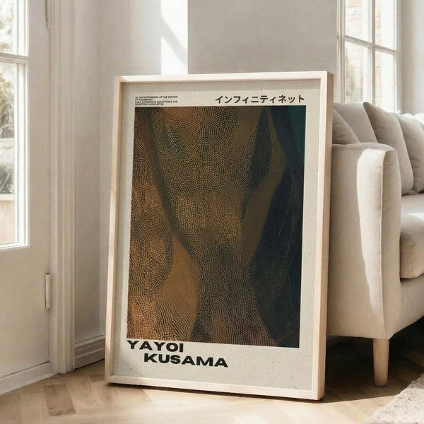 May include: Framed art print leaning against white wall next to beige upholstered chair in bright living room with large windows. Print features abstract dotted pattern in earth tones of brown, gold and dark green with Japanese text at top and "YAYOI KUSAMA" text at bottom. Natural wood frame with white matting, approximately 2-3 feet tall, positioned on light wooden floor.