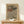 May include: Framed art print with wavy, flowing lines in earth tones of brown, beige, and muted colors displayed on a light wood table against a white wall. A small white wrapped gift with greenery sits beside the wooden frame. The artwork features organic, swirling patterns reminiscent of topographical maps or natural formations.