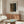 May include: Modern living room with cream walls featuring a framed exhibition poster displaying abstract dotted patterns in brown and blue tones. The poster hangs above a mid-century walnut credenza with brass hardware. A white curved chair sits nearby alongside a circular glass coffee table with colorful abstract art underneath. Minimalist copper floor and table lamps complete the contemporary Scandinavian-inspired setting.