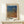 May include: Framed art poster displayed on light wood surface against cream wall. The poster features abstract dotted pattern in blue, orange and brown tones resembling flowing fabric or nets. Japanese text appears at top, artist name at bottom. Small white gift box with greenery sits beside the natural wood frame. Minimalist Scandinavian-style interior setting with soft natural lighting.