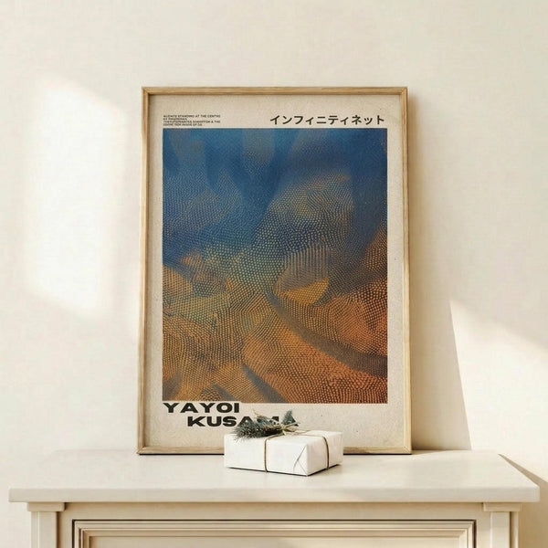 May include: Framed art poster displayed on light wood surface against cream wall. The poster features abstract dotted pattern in blue, orange and brown tones resembling flowing fabric or nets. Japanese text appears at top, artist name at bottom. Small white gift box with greenery sits beside the natural wood frame. Minimalist Scandinavian-style interior setting with soft natural lighting.