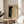 May include: Framed art print with black and cream polka dot pattern creating an infinity net design, displayed on a round wooden side table next to a beige upholstered chair. Natural sunlight streams across the neutral-toned room, casting soft shadows on the white walls. The artwork features dense dots transitioning from black to light areas, creating dimensional depth and movement within a thin wooden frame.