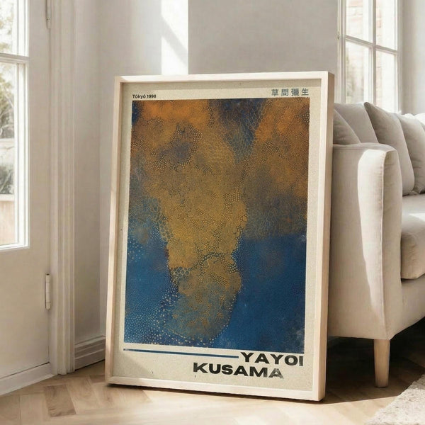 May include: A framed art print leaning against a white wall next to a beige sofa in a bright room with large windows. The poster features an abstract composition of golden yellow and deep blue dotted patterns that appear to flow and merge together. Text reading "YAYOI KUSAMA" appears at the bottom along with "Tokyo 1998" and Japanese characters at the top. The artwork is displayed in a natural wood frame on light wooden flooring.