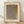 May include: Framed art print leaning against white wall on wooden floor. Cream-colored poster features abstract dotted pattern in brown and gray tones with organic flowing shapes. Text reads "YAYOI KUSAMA" at top and Japanese characters with "infinity nets" at bottom. Natural wood frame with light shadows cast across scene from window light.