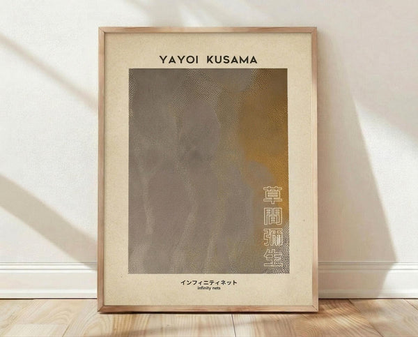 May include: Framed art print leaning against white wall on wooden floor. Cream-colored poster features abstract dotted pattern in brown and gray tones with organic flowing shapes. Text reads "YAYOI KUSAMA" at top and Japanese characters with "infinity nets" at bottom. Natural wood frame with light shadows cast across scene from window light.