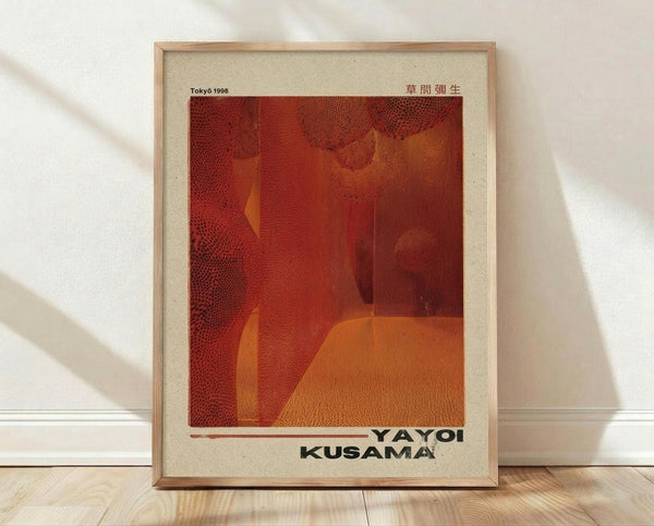 May include: Framed art print leaning against white wall on light wood floor. Poster features abstract red and orange dotted pattern with organic shapes and polka dot textures characteristic of contemporary Japanese art. Black text at bottom reads "YAYOI KUSAMA" with "Tokyo 1998" at top and Japanese characters in corner. Natural wood frame with cream matting, soft window light casting shadows across scene.