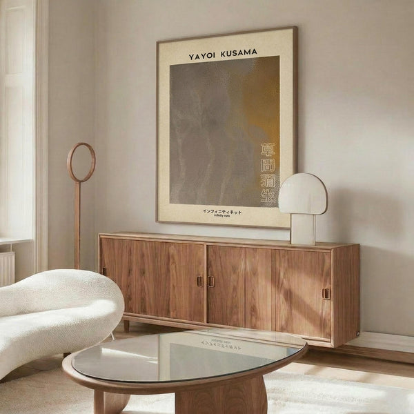 May include: Minimalist living room featuring a framed abstract poster with neutral tones and Japanese text mounted above a wooden credenza. The poster displays textured dotted patterns in muted grays and golds. Modern furniture includes a curved white sculptural chair, round glass coffee table, and copper floor lamp, creating a warm contemporary interior scene.