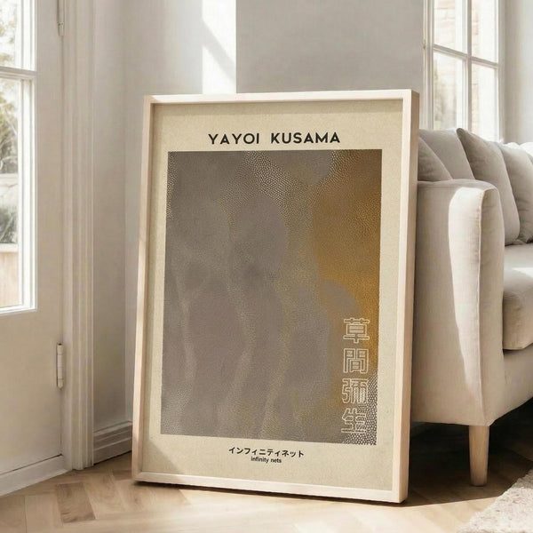 May include: Framed art print leaning against white wall next to beige sofa in bright living room. The poster features abstract dotted pattern in brown and gold tones with "YAYOI KUSAMA" text at top and Japanese characters vertically aligned on right side. Light wood frame with cream matting, approximately 2-3 feet tall, positioned on hardwood floor with natural sunlight streaming through windows.