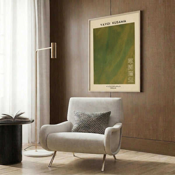 May include: Framed Yayoi Kusama exhibition poster with green textured artwork and Japanese text, displayed above a cream upholstered chair with geometric patterned pillow. Modern reading nook with brass floor lamp, dark round side table with open book, wood panel wall, and natural lighting from window with curtains.