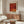 May include: Framed art print with abstract red and orange composition hanging above mid-century walnut credenza in minimalist living room. White upholstered chair and glass coffee table visible. Poster features geometric shapes in warm tones with Japanese text at top and artist name at bottom. Natural lighting creates shadows across polished wood surfaces.