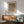 May include: Framed Yayoi Kusama poster with dotted pattern artwork in neutral tones, displayed above wooden sideboard in modern living room setting. Green upholstered armchair with wooden frame sits nearby, white ceramic table lamp on credenza. Natural sunlight creates shadows across white walls, showing minimalist Scandinavian-style interior design with warm wood furniture and clean aesthetic.
