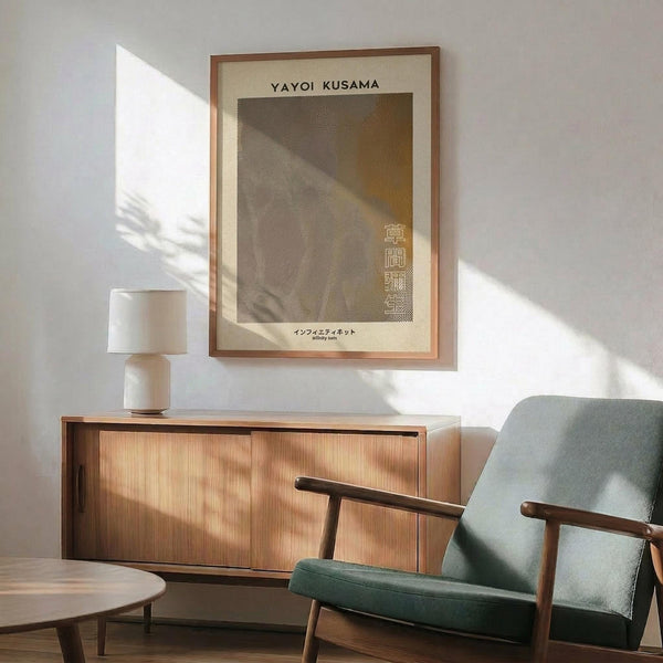 May include: Framed Yayoi Kusama poster with dotted pattern artwork in neutral tones, displayed above wooden sideboard in modern living room setting. Green upholstered armchair with wooden frame sits nearby, white ceramic table lamp on credenza. Natural sunlight creates shadows across white walls, showing minimalist Scandinavian-style interior design with warm wood furniture and clean aesthetic.