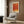 May include: Orange and red abstract art poster with Japanese text and "Yayoi Kusama" displayed on wood paneled wall above beige upholstered armchair with geometric patterned pillow. Modern brass floor lamp and dark side table with open book visible. Warm neutral interior with natural lighting from window.