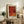 May include: Red and orange abstract art poster in light wood frame displayed on rustic wooden console table alongside fern plant, cream ceramic vase, and stack of books. Warm neutral living room setting with beige armchair visible in foreground.