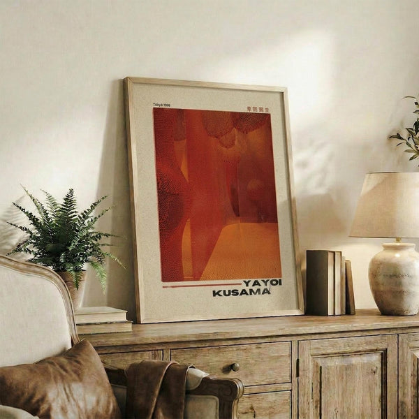 May include: Red and orange abstract art poster in light wood frame displayed on rustic wooden console table alongside fern plant, cream ceramic vase, and stack of books. Warm neutral living room setting with beige armchair visible in foreground.