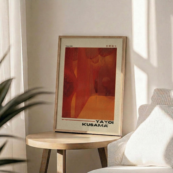 May include: Framed art print with red and orange abstract design featuring "YAYOI KUSAMA" text at bottom, displayed on round wooden side table next to beige upholstered chair. Natural lighting creates soft shadows across white walls. Green plant leaves visible in corner. Minimalist Scandinavian-style interior setting with neutral tones.