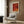 May include: A framed vintage-style poster with red and orange tones featuring abstract curved forms, displayed on wood paneled wall above a cream upholstered chair with geometric patterned pillow. Modern floor lamp with brass finish stands nearby next to small dark round table with open book. Warm neutral interior with natural lighting from window.