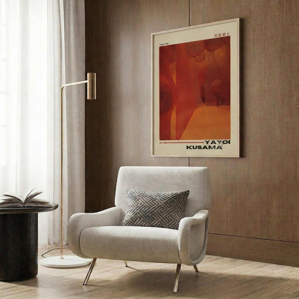 May include: A framed vintage-style poster with red and orange tones featuring abstract curved forms, displayed on wood paneled wall above a cream upholstered chair with geometric patterned pillow. Modern floor lamp with brass finish stands nearby next to small dark round table with open book. Warm neutral interior with natural lighting from window.