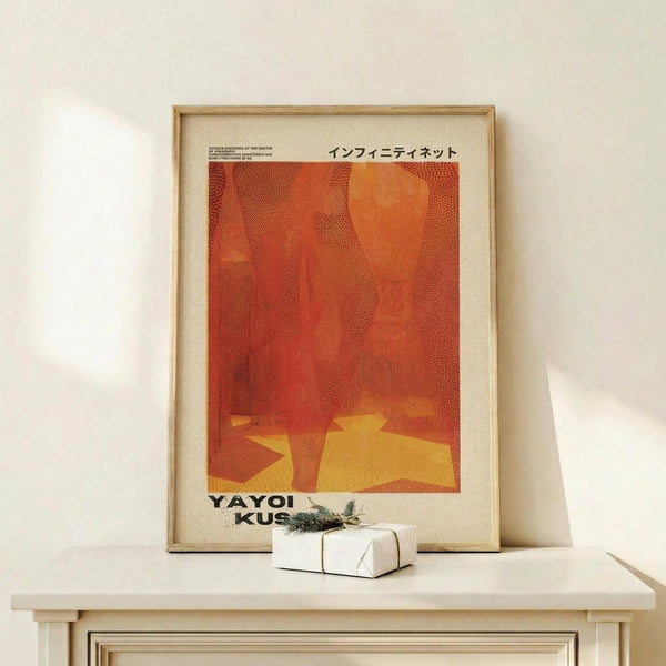 May include: Framed vintage-style art poster with abstract orange and red dotted pattern, displayed on light wood surface alongside small white wrapped gift with evergreen sprig, set against cream wall in minimalist interior setting.
