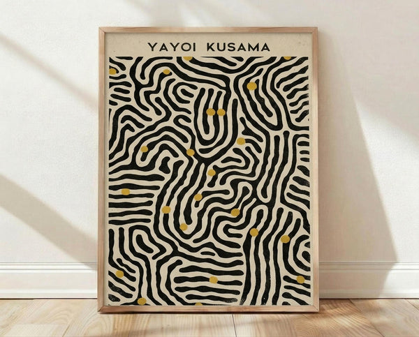 May include: A framed art print with black wavy lines creating an abstract maze-like pattern on cream background, accented with small golden yellow dots scattered throughout. The print displays "YAYOI KUSAMA" text at the top and is shown in a light wood frame, leaning against a white wall on wooden flooring with natural lighting and shadows.