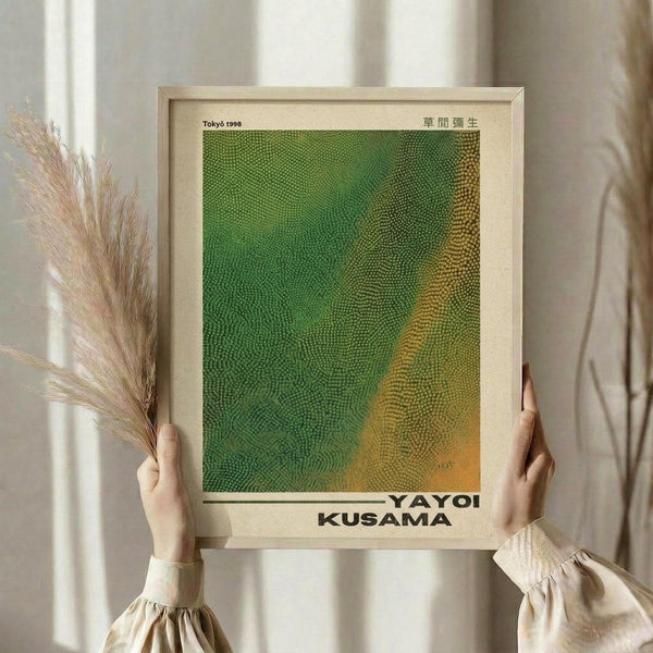 May include: Person in cream ruffled blouse holding framed art print in light wooden frame. Print features green and orange gradient dotted pattern reminiscent of organic textures. Person also holds dried wheat or pampas grass stems. Soft natural lighting against neutral curtained background creates minimalist aesthetic.
