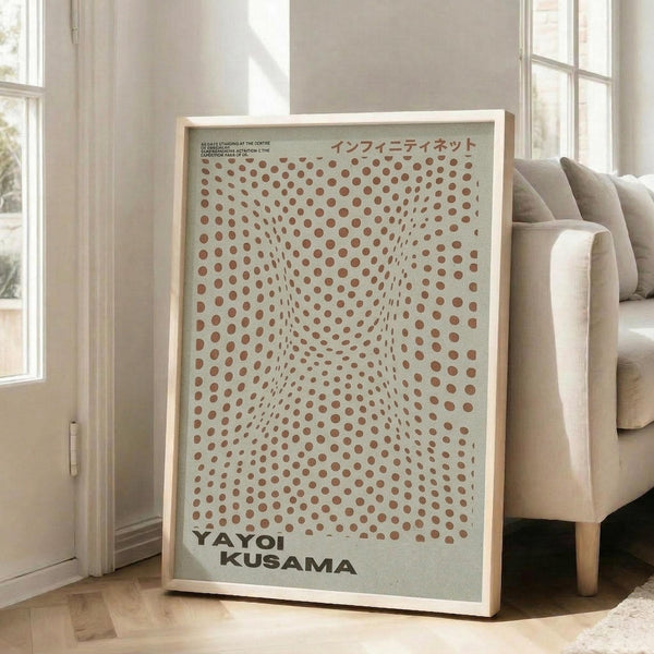 May include: Framed art print with cream background and reddish-brown polka dots creating an optical illusion of depth and movement, featuring Japanese text at top and "YAYOI KUSAMA" at bottom in black letters. Print displayed in light wood frame leaning against white wall next to beige upholstered sofa in bright, naturally-lit room with white walls and windows.