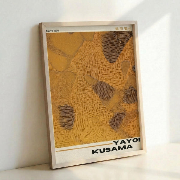 May include: Framed poster with yellow and orange dotted pattern artwork leaning against white wall. The print features Kusama's signature polka dot style in warm golden tones with organic flowing shapes. Black text at bottom reads "YAYOI KUSAMA" with "TOKYO 1998" in upper corner. Light wooden frame surrounds the contemporary art print in minimalist room setting.