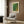 May include: Modern cream armchair with geometric throw pillow in contemporary living room setting with wood-paneled walls. Green and yellow abstract dotted art poster in light wooden frame hangs above chair. Gold floor lamp and dark side table with open book complete the minimalist interior design scene.