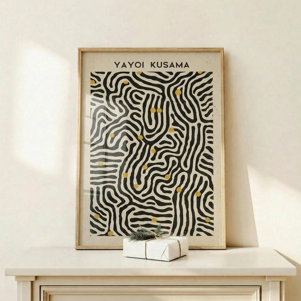 May include: Framed abstract art print with flowing black wavy lines and small yellow dots on cream background, displayed on light wood console table beside white wrapped gift with green sprig. Thin natural wood frame against white wall in minimalist room setting.