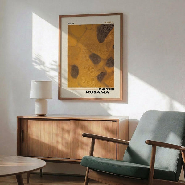 May include: Framed Yayoi Kusama inspired art print with yellow and orange dotted pattern and dark organic shapes, displayed above a wooden mid-century modern credenza with white ceramic lamp. Green upholstered armchair with wooden frame sits nearby in bright, sunlit room with white walls and wooden coffee table.