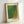 May include: Framed art print leaning against white wall showing abstract green and yellow dotted pattern resembling reptile skin texture. Artwork features "YAYOI KUSAMA" text at bottom with "Tokyo 1998" and Japanese characters at top. Natural wood frame with cream matting displays the geometric halftone dot design that transitions from bright green on left to golden yellow on right.