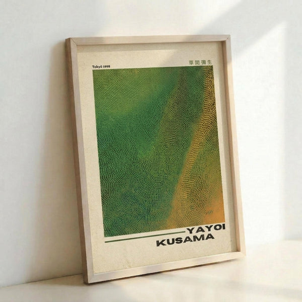 May include: Framed art print leaning against white wall showing abstract green and yellow dotted pattern resembling reptile skin texture. Artwork features "YAYOI KUSAMA" text at bottom with "Tokyo 1998" and Japanese characters at top. Natural wood frame with cream matting displays the geometric halftone dot design that transitions from bright green on left to golden yellow on right.
