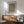 May include: Framed art print with black and yellow wavy line pattern hanging above mid-century modern wooden sideboard. Green upholstered armchair with wooden frame sits beside credenza. White ceramic table lamp and round coffee table visible. Warm natural light creates shadows on white wall in minimalist living room setting.