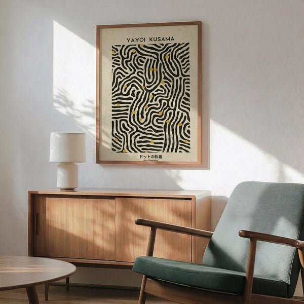 May include: Framed art print with black and yellow wavy line pattern hanging above mid-century modern wooden sideboard. Green upholstered armchair with wooden frame sits beside credenza. White ceramic table lamp and round coffee table visible. Warm natural light creates shadows on white wall in minimalist living room setting.