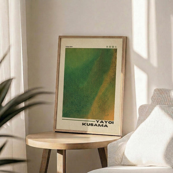 May include: Framed art print featuring green and yellow dotted pattern in wood frame, displayed on round wooden side table next to beige upholstered chair with white throw pillow. Natural sunlight creates soft shadows on white walls, with plant leaves visible in corner of bright, minimalist living room setting.