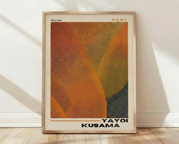 May include: Framed art print featuring abstract organic shapes in warm orange, yellow, and dark blue-green tones with halftone dot pattern texture. Text reads "Tokyo 1998" and "YAYOI KUSAMA" with Japanese characters. Light wood frame displayed against white wall on wooden floor with natural lighting creating soft shadows.