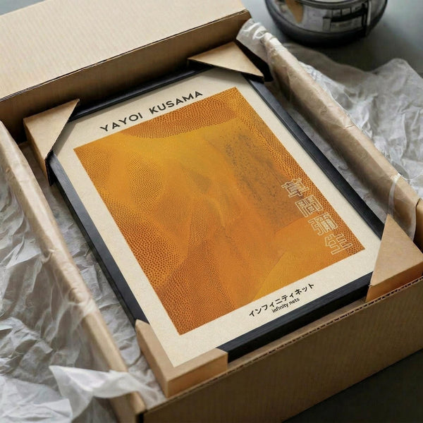 May include: Orange and yellow gradient artwork with dotted pattern texture displayed in black frame, packaged in brown cardboard box with white tissue paper. Two wooden dowels visible alongside frame. Warm amber glass vessel partially shown in background on neutral surface.