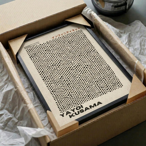 May include: Framed beige poster with black polka dot pattern in halftone style, displayed in black frame inside cardboard shipping box with protective tissue paper. Wooden dowels visible alongside for hanging. Clean minimalist design with dense dots creating gradient effect from top to bottom.