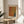 May include: Framed art print with abstract orange, yellow, green and blue dotted pattern resembling flowing fabric or waves, displayed on a light wooden round side table next to a beige upholstered chair. Natural sunlight streams across white walls creating soft shadows, with green plant leaves visible in corner. The artwork features a cream colored mat and thin wooden frame.