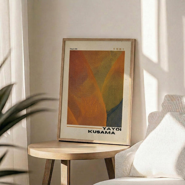 May include: Framed art print with abstract orange, yellow, green and blue dotted pattern resembling flowing fabric or waves, displayed on a light wooden round side table next to a beige upholstered chair. Natural sunlight streams across white walls creating soft shadows, with green plant leaves visible in corner. The artwork features a cream colored mat and thin wooden frame.