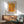 May include: Framed Yayoi Kusama Infinity Nets art print in warm golden orange tones with Japanese text, displayed above a mid-century modern wood credenza with white ceramic lamp. Mint green upholstered armchair with wooden frame sits beside round wood coffee table. Bright natural sunlight creates geometric shadows across white wall in minimalist living room setting.
